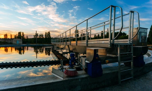 Spokojny wieczór na nabrzeżu zbiornika wodnego, gdzie promienie słońca odbijają się od wody, a poręcz prowadzi drogę. Zachód słońca rozświetla wodę i ogrodzenie ciepłym blaskiem.